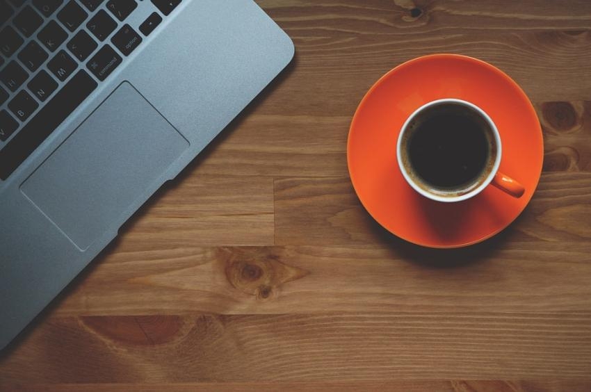 Rote Tasse mit Kaffee auf Unterteller steht auf einer Holzplatte, links davon ist ein Touchpad mit Tastatur