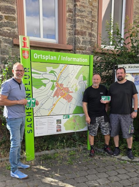 Drei Personen stehen an einer Infotafel mit einem Ortsplan von Sachsenflur